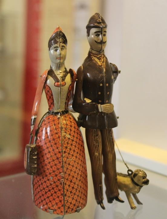 Eine Frau im langen Kleid, ein Mann im Anzug und ihr Mops an der Leine: Ehepaar Lehmann als Blechspielzeug (um 1900)