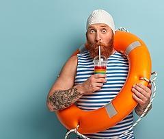 Embarrassed holiday maker drinks cold summer cocktail, spends free time at beach, wears swimcap sailor t shirt, swims with lifebuoy, has surprised expression, models over blue wall with free space