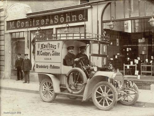 Ein altertümlicher Lieferwagen (um 1910) mit der Aufschrift M. Conitzer und Söhne mit zwei Angestellten, im Hintergrund das Warenhaus