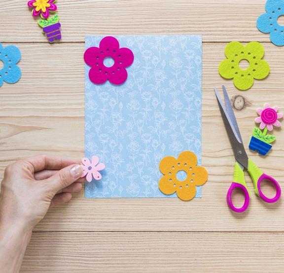 Buntes Bastelmaterial: Filzblumen, Karte, Schere, eine Hand hält eine Filzblume