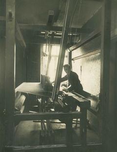 Ein Mann steht an einem Webstuhl in einem sehr kleinen Raum. Es ist der letzte Webstuhl dieser Art, das Foto wurde 1925 aufgenommen.