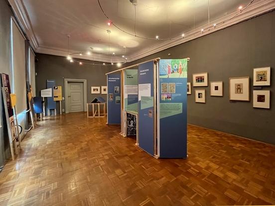 Im Saal stehen in der Mitte Ausstellungstafeln in kräftigen Farben, rechts an der grauen Wand hängen die Originalbilder von Paul Goesch.