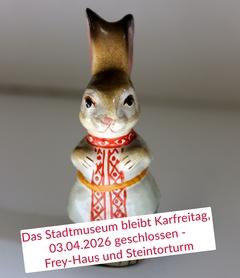 Aufrecht stehender Hase mit weißem, musterverziertem Gewand aus glasierter Keramik, dazu die Infotafel zur Schließzeit zum Karfreitag 2026
